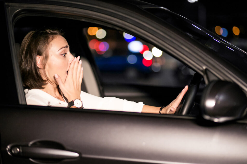 Femme assise au volant d'une voiture à l'arrêt, regard tendu, main sur la bouche signe de panique, illustrant l'amaxophobie et la peur de conduire au volant.