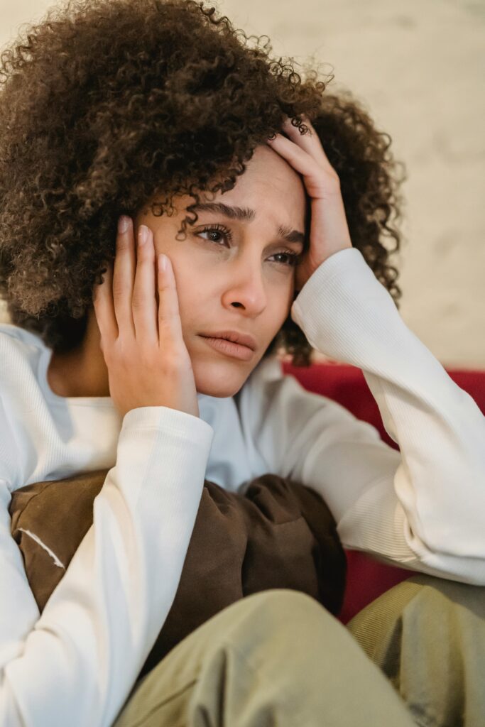 Despaired African American female with curly hair touching face and looking away with sorrow while sitting on sofa at home