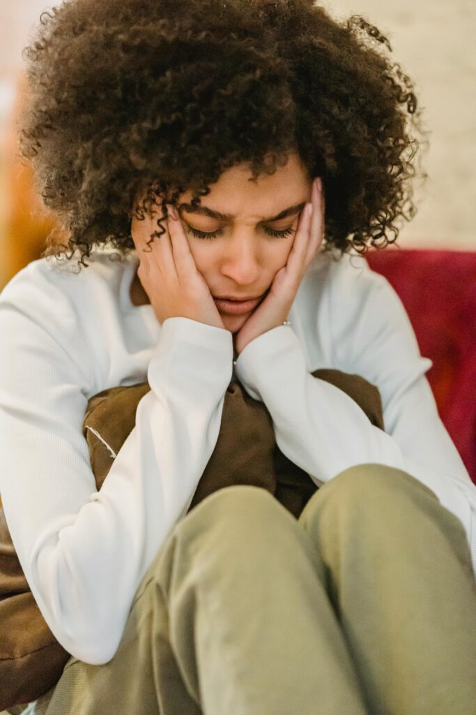 Young sad ethnic lady in casual outfit sitting with pillow with hands on head and closed eyes in light room